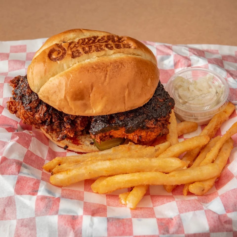 A Student Burger spicy chicken sandwich with pickles, fries, and coleslaw on checkered paper.