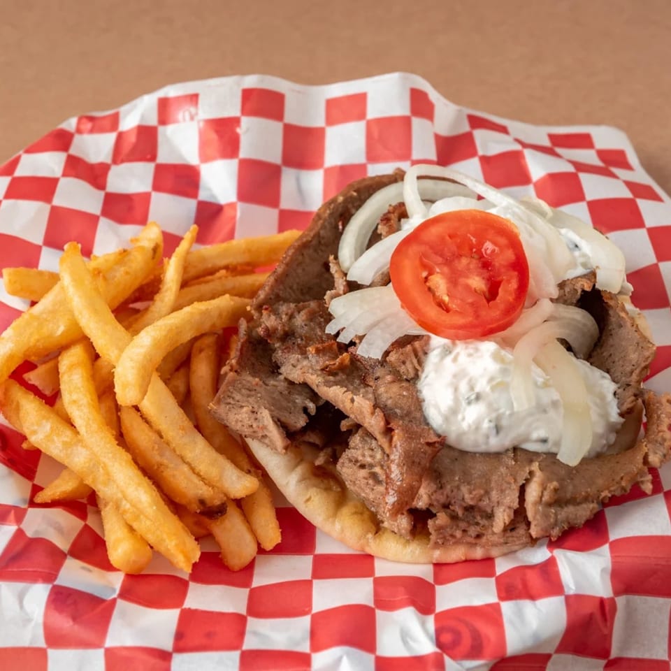 A gyro with sliced meat, onions, tomato, and white sauce, served with golden fries on checkered paper.