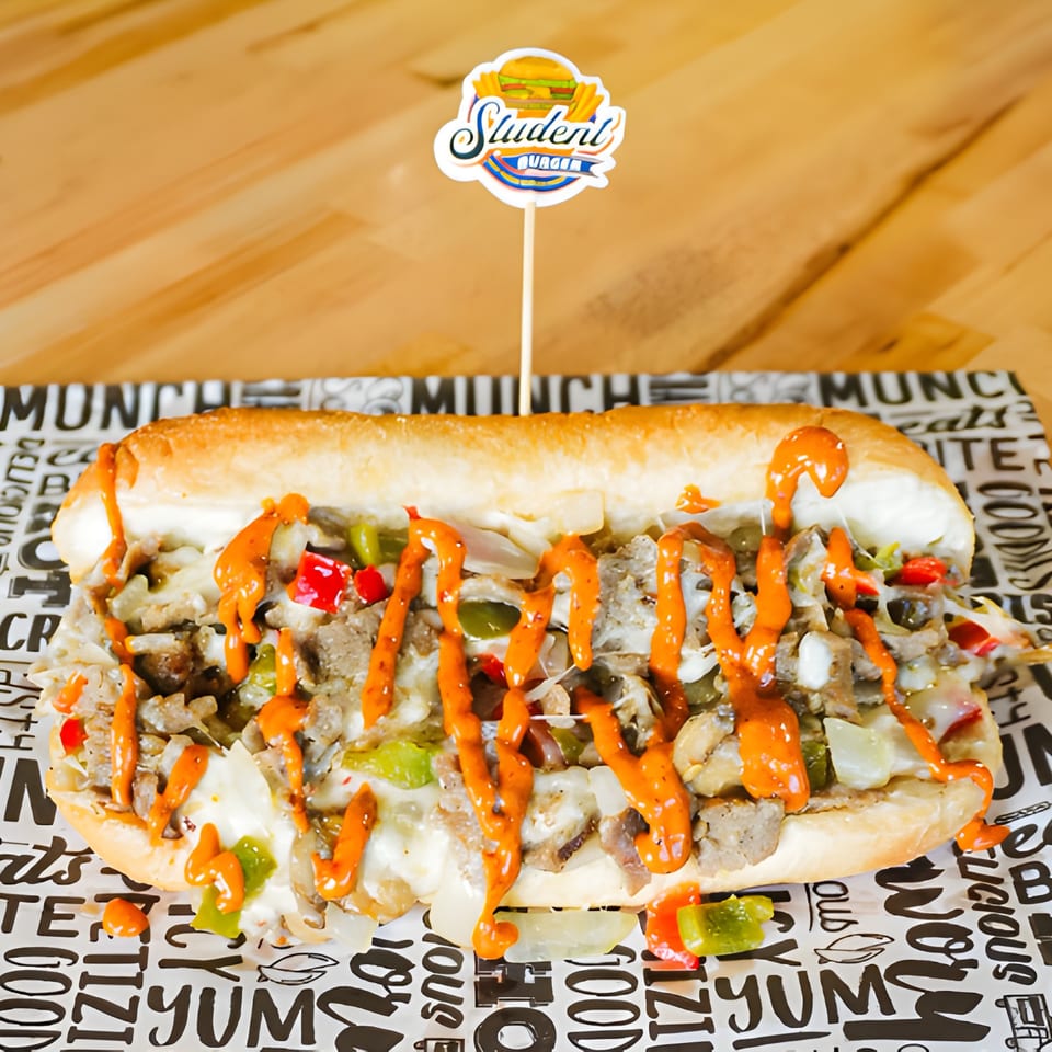 A Student Burger cheesesteak sandwich with orange sauce, peppers, and onions on branded paper.